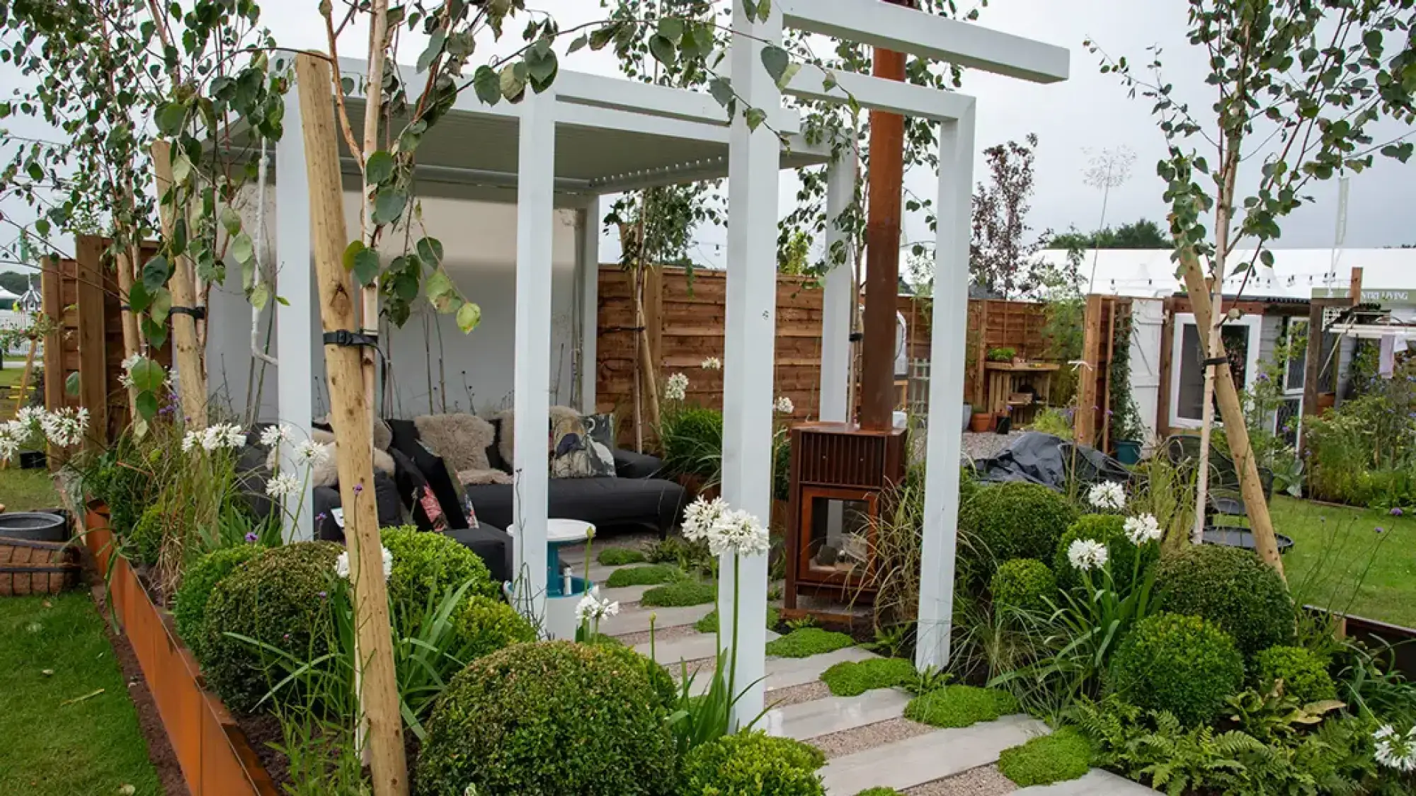 A contemporary terrace garden with a white pergola, grey sofa, corten steel fireplace, and stepping stone path through green plants, white flowers.