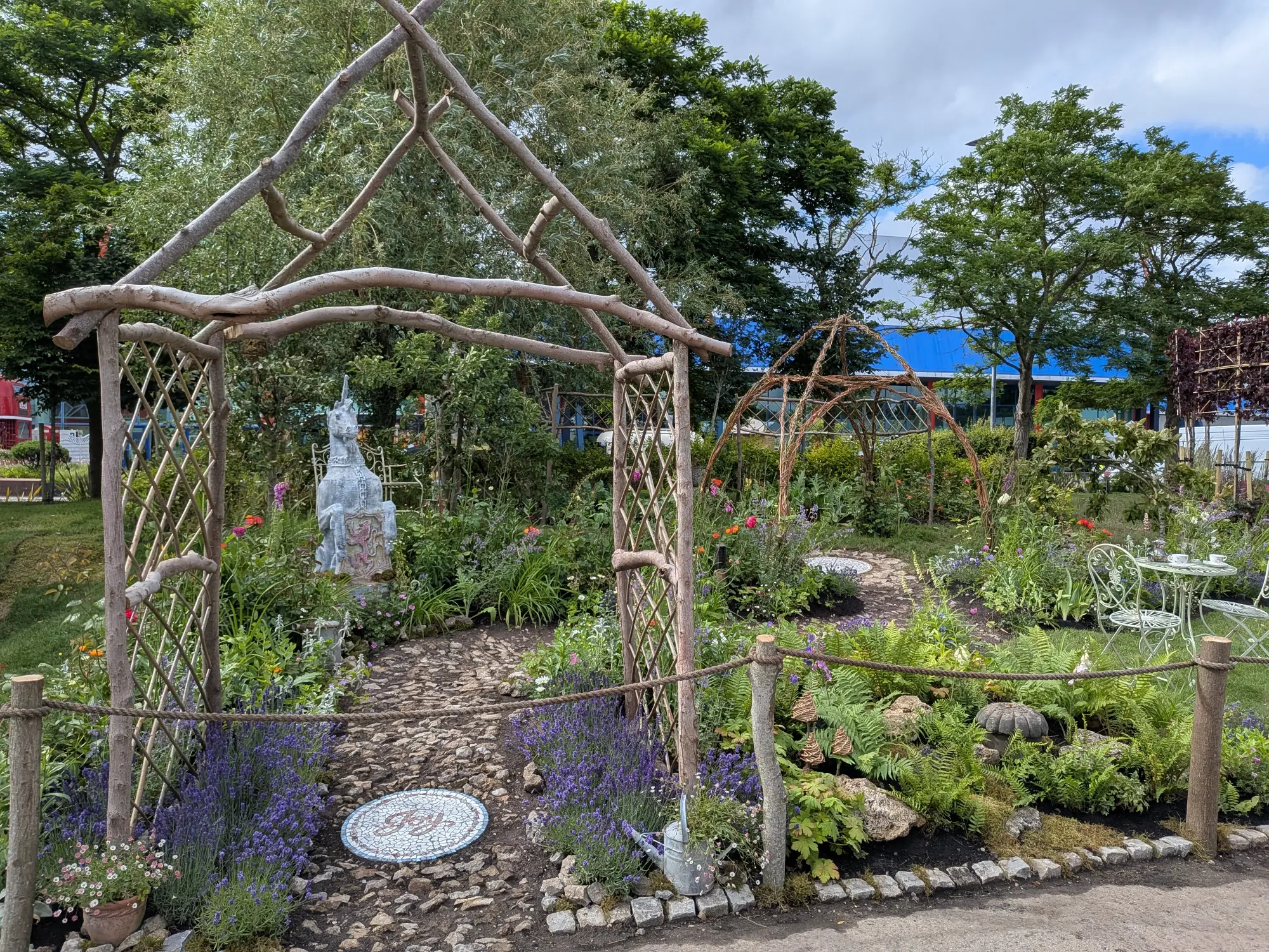 A whimsical show garden with a woven wood archway, a cobblestone path, a white unicorn sculpture, and a seating area surrounded by lavender and mixed perennials.