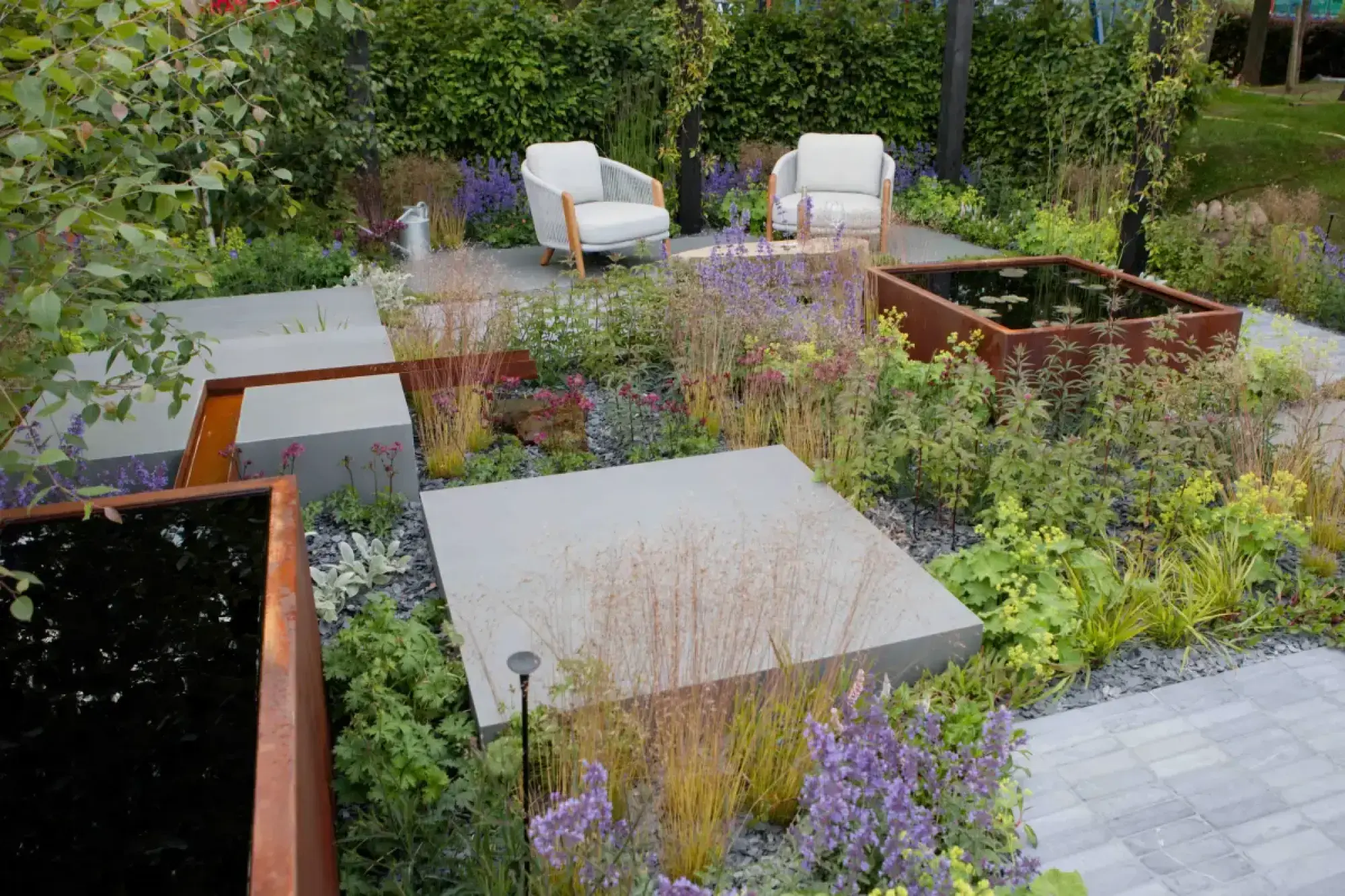 A contemporary garden features rust-coloured corten steel water features, grey paving, and naturalistic plantings of ornamental grasses and purple flowers, with two light outdoor armchairs.