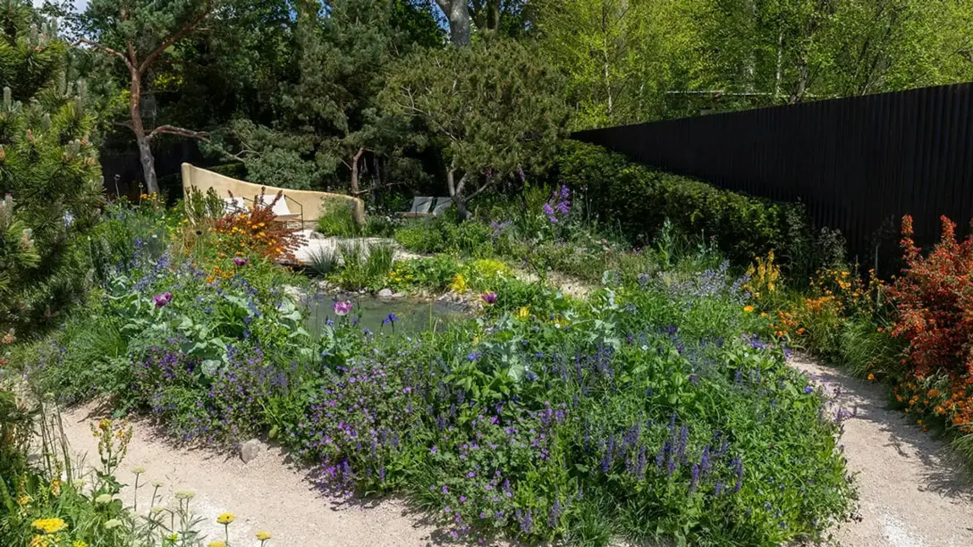 A naturalistic garden featuring a curved beige bench, a pond, winding gravel paths, and abundant planting with purple, blue, yellow, and orange flowers.