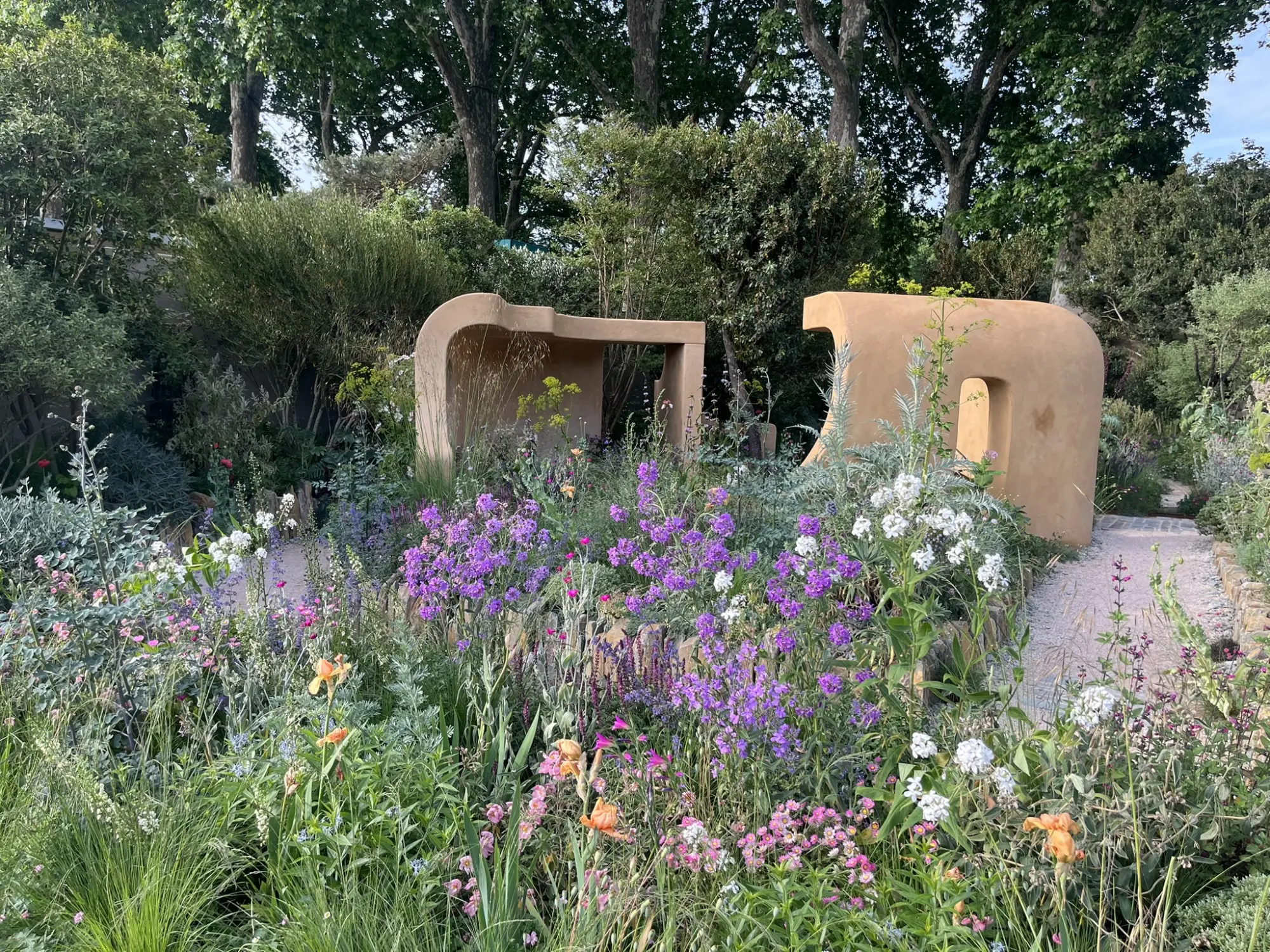 A show garden with two curved, earth-toned structures, abundant meadow planting with purple and pink flowers, and a gravel path.