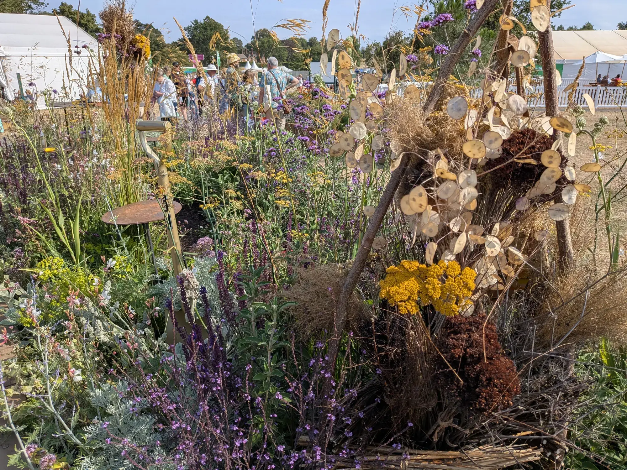 A long border garden featuring a rustic branch sculpture with dried Lunaria pods, abundant naturalistic planting of purple verbena, yellow yarrow, and silver foliage, and a leaning garden spade.