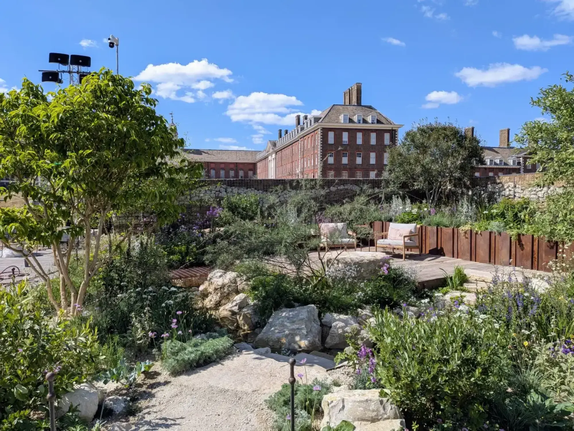 A small naturalistic garden has a Corten steel wall, large pale boulders, a timber deck with two chairs, and mixed green, purple, and white planting.