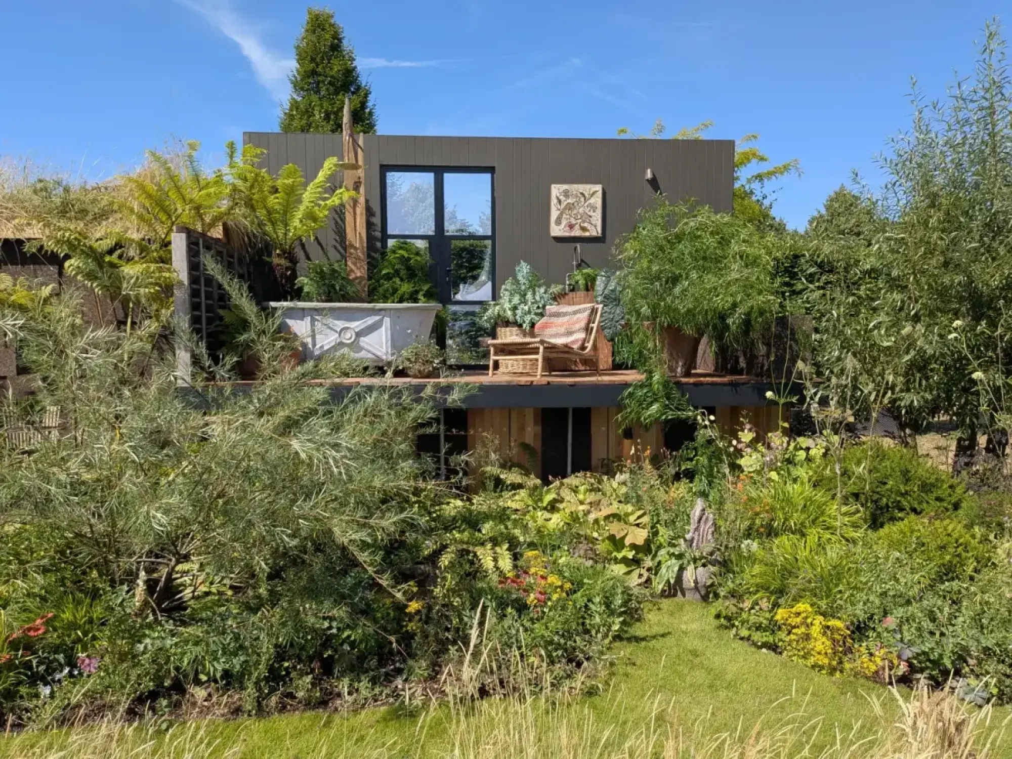 A modern balcony garden features an outdoor bathtub, a dark grey facade, wooden decking, a rattan chair, and lush green planting.