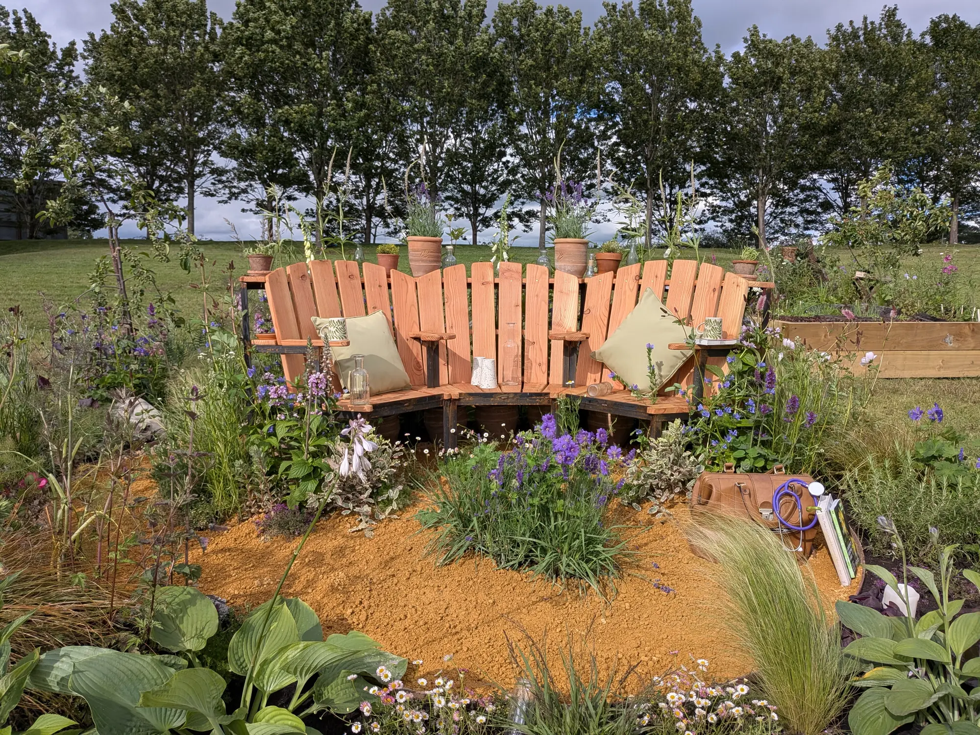 A U-shaped wooden bench with cushions sits around a sandy path blooming with purple and white flowers, grasses, and hostas, with a doctor's bag.