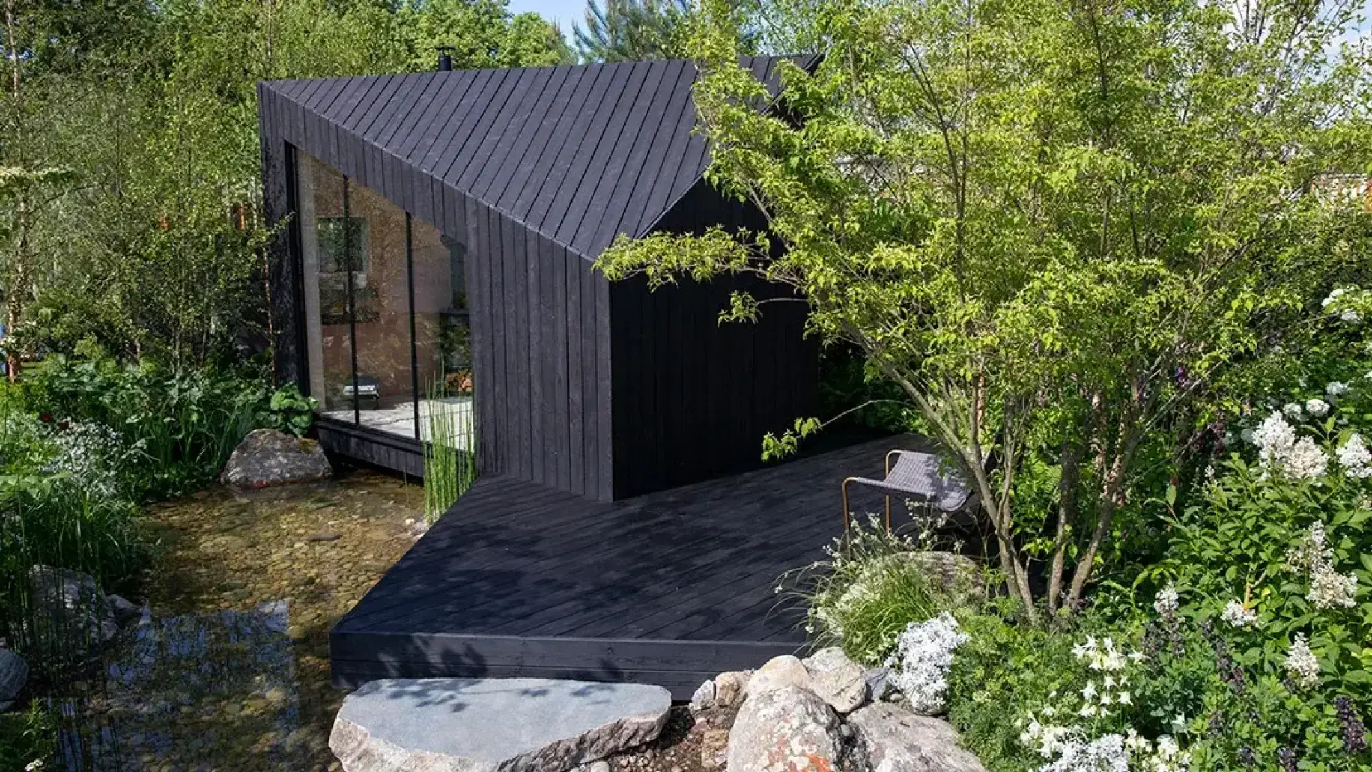 A modern black timber cabin with a sharp angled roof and glass wall overlooks a shallow pebble stream, surrounded by lush green foliage and white flowers.