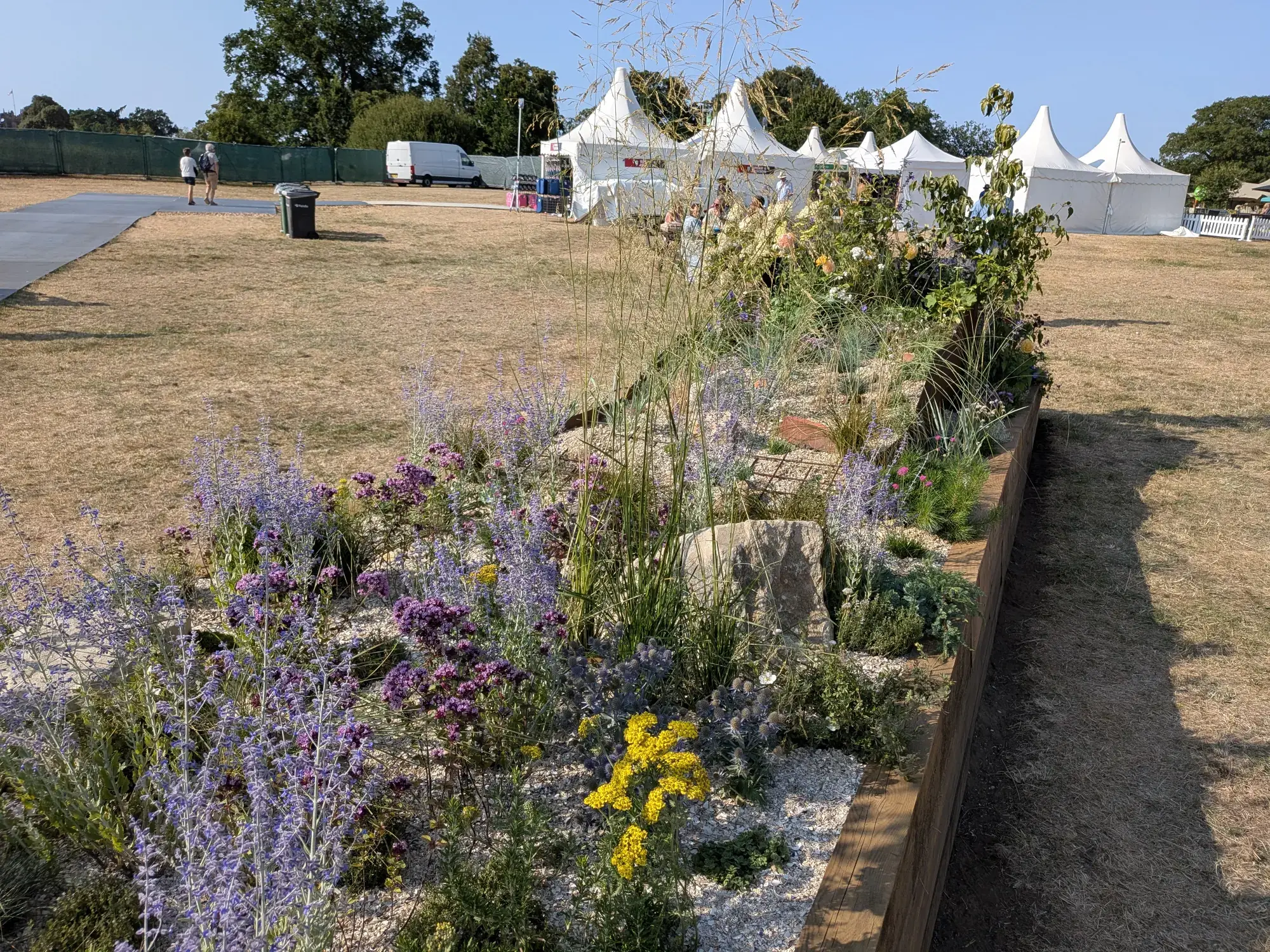 A long, timber-edged raised border displays an informal array of purple, yellow, and blue flowers, grasses, rocks, and gravel.