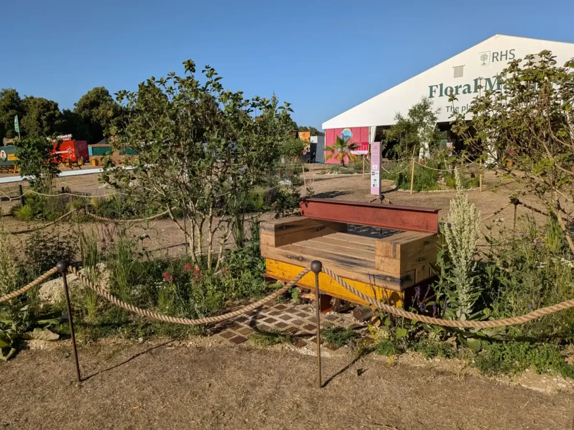 A small pocket planting garden featuring a reclaimed timber bench with a yellow base and red metal beam, surrounded by mixed herbaceous plants and small trees.