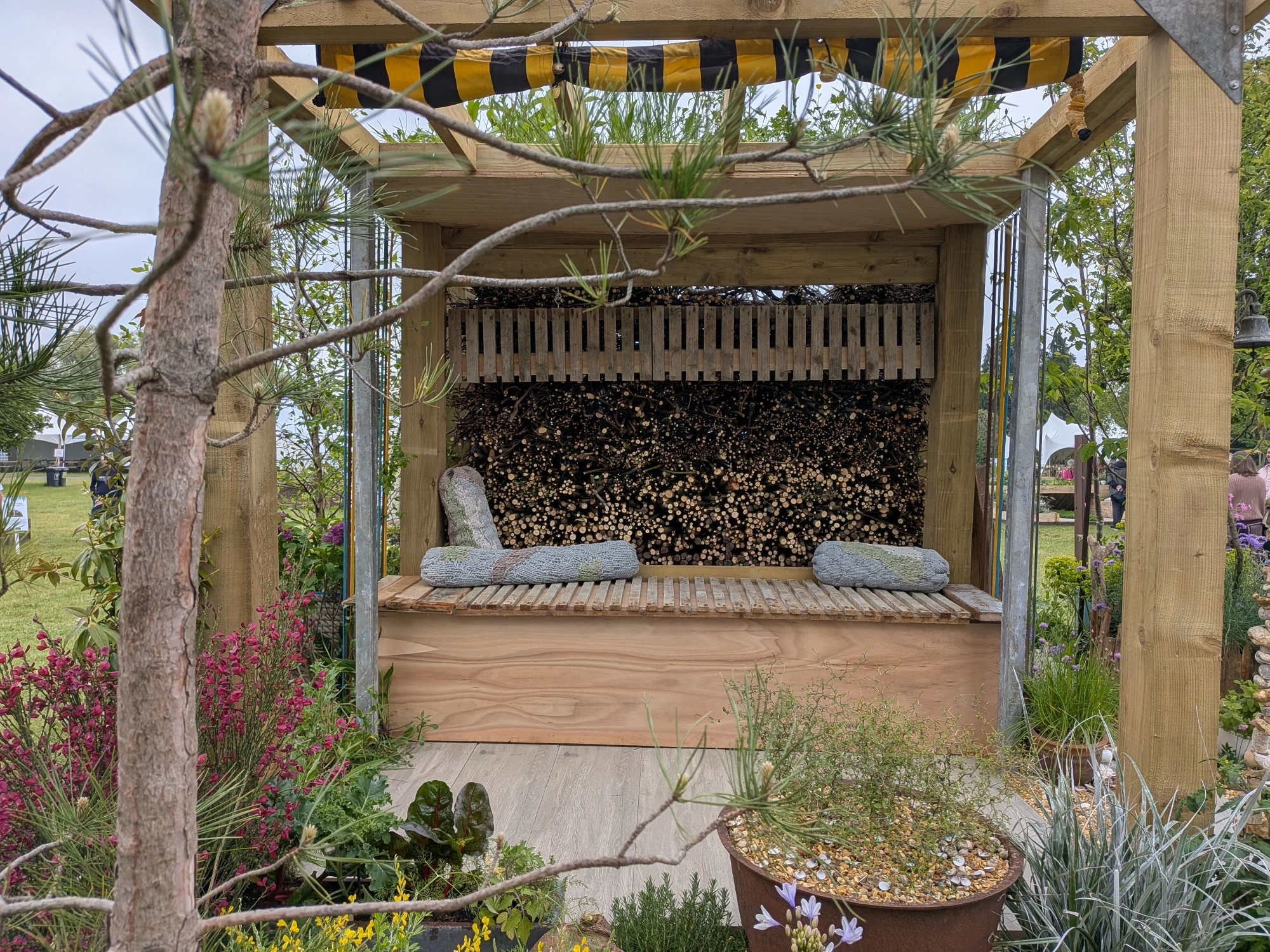 A wooden shelter offers a slatted bench with knitted cushions, backed by a large stacked log and timber bug hotel, surrounded by pine and colourful plants.